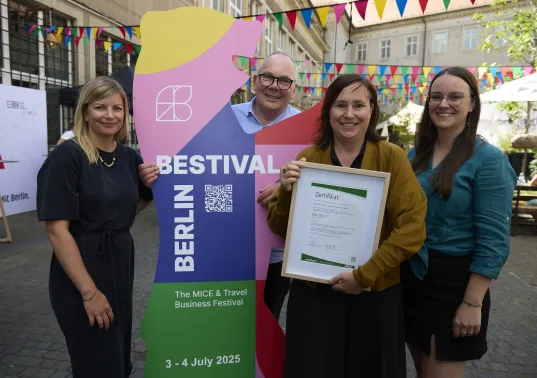 Vier lächelnde Personen posieren draußen mit einer Urkunde vor einem bunten Festival-Schild.