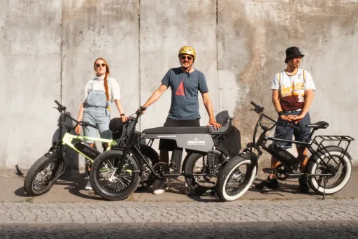 Drei Personen mit E-Bikes stehen vor einer Betonwand in der Sonne.