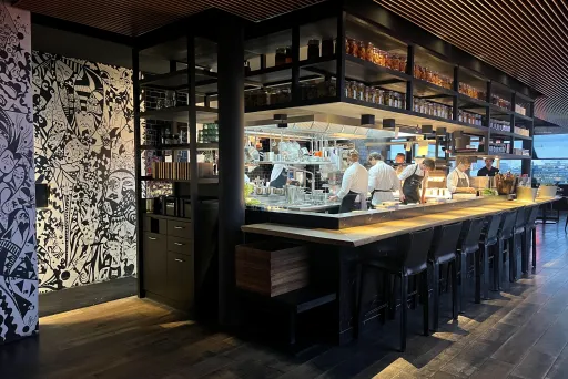 Moderne Bar mit offenen Regalen, Köche arbeiten hinter dem Tresen, gedämpftes Licht.