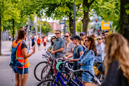 Gruppe von Radfahrer*innen hört einer Guide auf einer sommerlichen Stadtstraße zu.