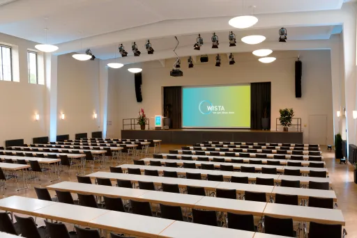 Großer, heller Konferenzsaal mit leeren Tischen und Leinwand auf der Bühne.
