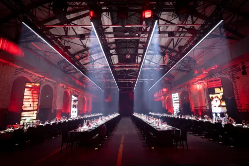 Langer Festsaal mit zwei gedeckten Tafeln, rote Beleuchtung, industrielle Atmosphäre.