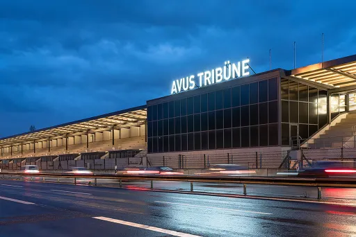 Stadiontribüne bei Dämmerung, beleuchtet, nasse Straße und vorbeifahrende Autos.