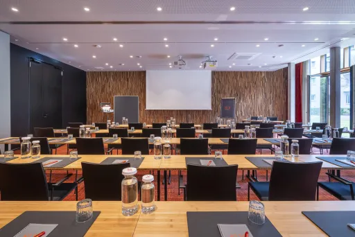Heller Konferenzraum mit Tischen, Stühlen, Wasserflaschen und Leinwand.