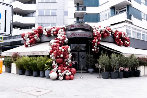 Restaurantfassade mit roter und weißer Luftballondekoration, umgeben von Pflanzen in Töpfen.