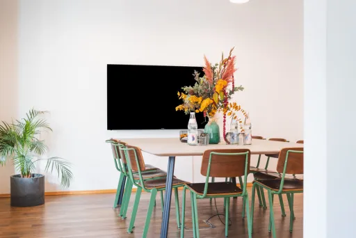 Sichtbar ist ein modern eingerichteter Meetingraum mit einem zentralen Tisch und 10 Stühlen. Der Raum ist hochwertig eingerichet mit einem Bildschirm und viel Helligkeit.