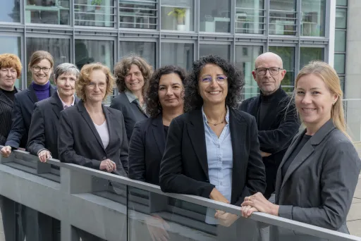 Neun Personen im Business-Outfit stehen lächelnd vor einem modernen Gebäude.