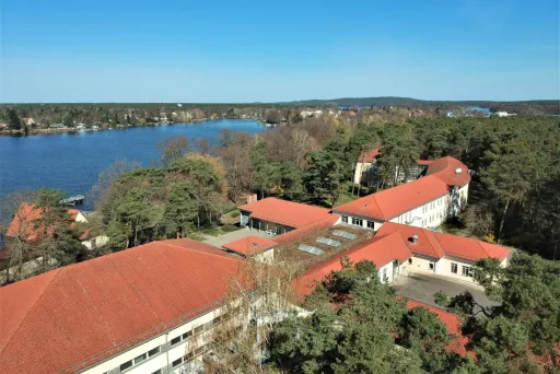 Gebäude mit roten Dächern am See, umgeben von Bäumen unter blauem Himmel.