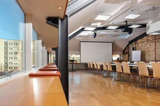 Heller Konferenzraum mit langen Tischen, Holzstühlen und großer Fensterfront