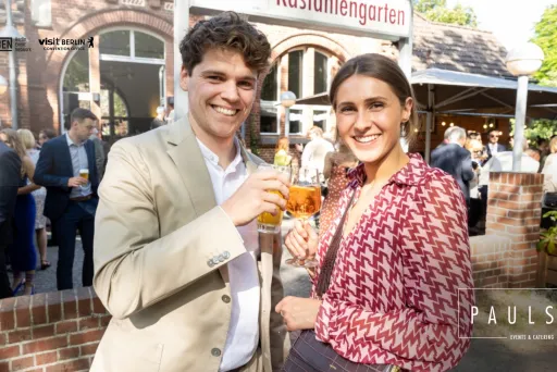Zwei lächelnde Personen stoßen im Freien mit Getränken an, im Hintergrund stehen weitere Gäste.