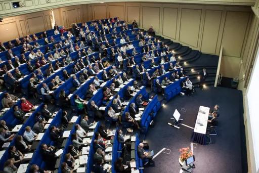 Hörsaal mit vielen Menschen auf blauen Sitzen, zwei Vortragende sitzen an Tisch