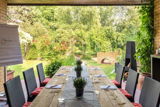 Langer Holztisch mit Stühlen auf Terrasse, Blick in grünen Garten.
