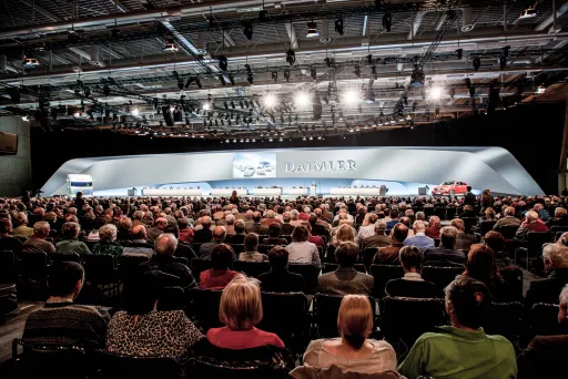 Großer Konferenzsaal mit vielen sitzenden Menschen vor einer beleuchteten Bühne.