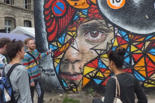 Bunte Graffiti-Wand mit Vogel- und Frauengesicht, mehrere Menschen davor.