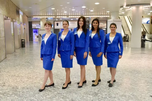 Fünf Flugbegleiterinnen in blauen Uniformen stehen in einer hellen Flughafenhalle.