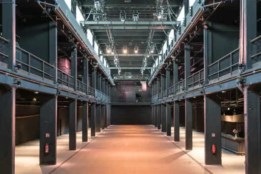 Große, leere Industriehalle mit Metallbalken und hoher Decke, Licht von oben.