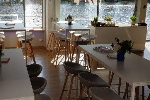 Helles Café mit weißen Tischen, Barhockern und Blick auf einen Fluss.