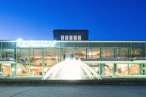 Modernes Gebäude mit großer Glasfront und beleuchtetem Springbrunnen am Abend.