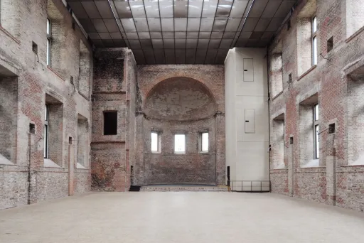 Große leere Halle mit Backsteinwänden und Glasdach, viel Tageslicht.