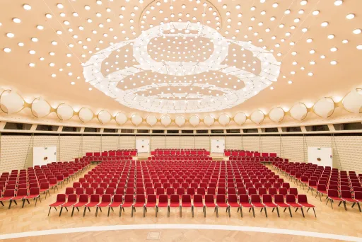 Großer leerer Saal mit roten Stühlen und eindrucksvoller Deckenbeleuchtung.