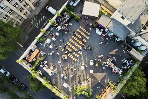 Dachterrasse mit Stühlen, Menschen und Tischen, umgeben von Gebäuden und Bäumen
