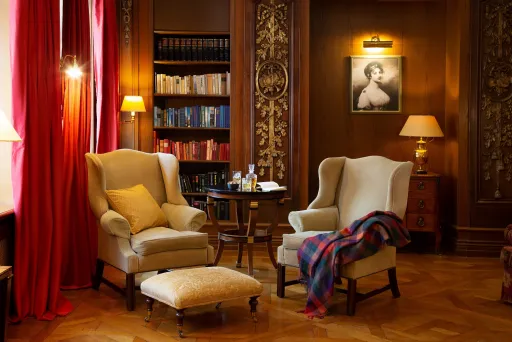 Gemütliche Bibliothek mit zwei Sesseln, rotem Vorhang, Bücherregal und warmem Licht.