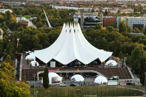 Weißes Zeltgebäude im Park, zahlreiche Besucher davor, Stadt im Hintergrund
