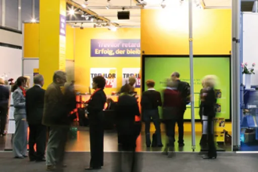 Menschen stehen und gehen vor einem gelb beleuchteten Messestand.