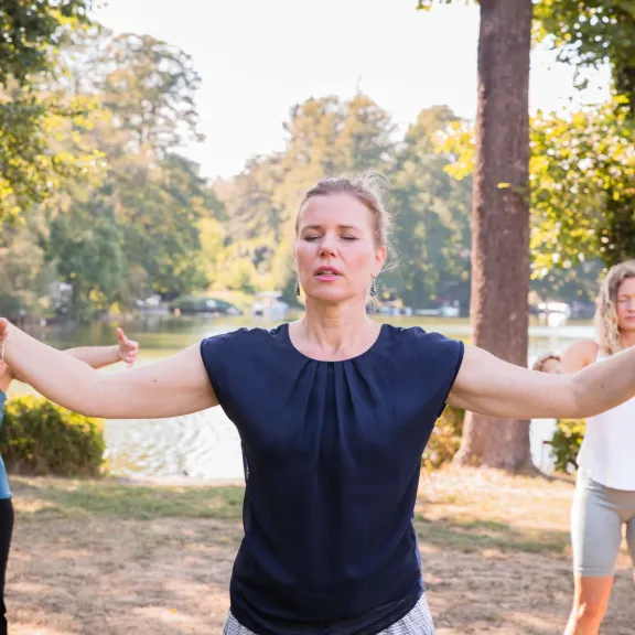 Drei Frauen machen Gymnastikübungen im Freien am See, umgeben von Bäumen.