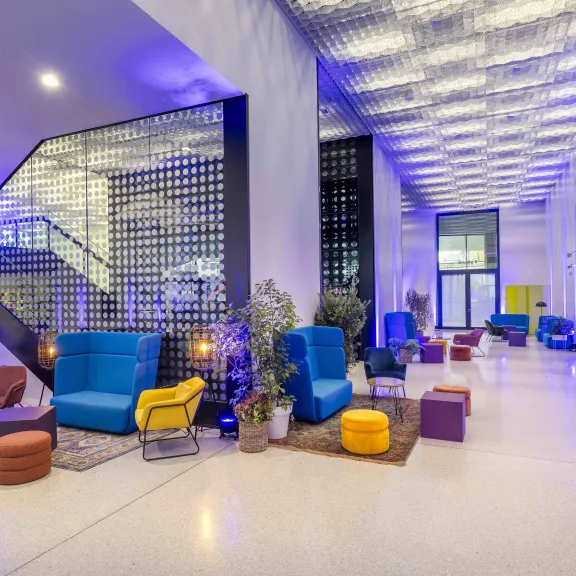 Modernes Foyer mit bunten Sitzmöbeln, Pflanzen und lila-blauer Beleuchtung.