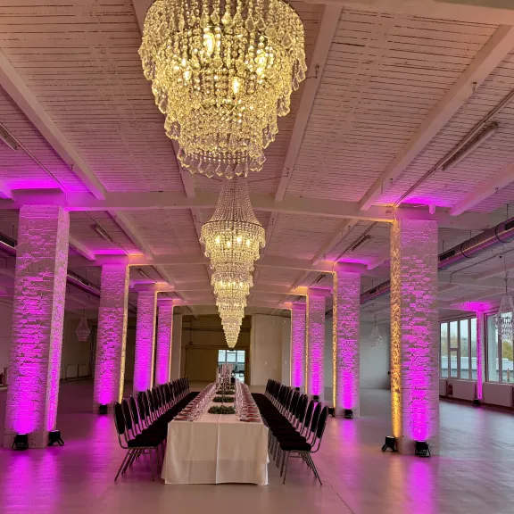 Langer Tisch mit schwarzen Stühlen, Kronleuchter und pink beleuchtete Säulen in Festsaal.