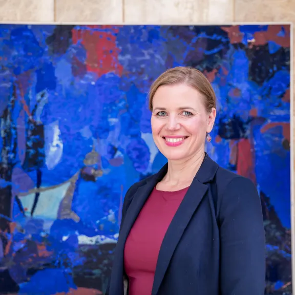 Frau mit Blazer lächelt vor abstraktem, blauem Gemälde.