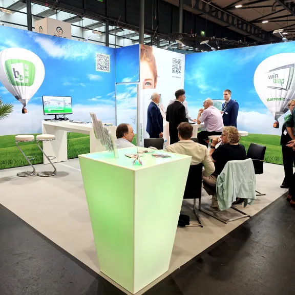 Messestand mit Menschen, grüner Theke, Stühlen und Himmel-Ballon-Motiv im Hintergrund.