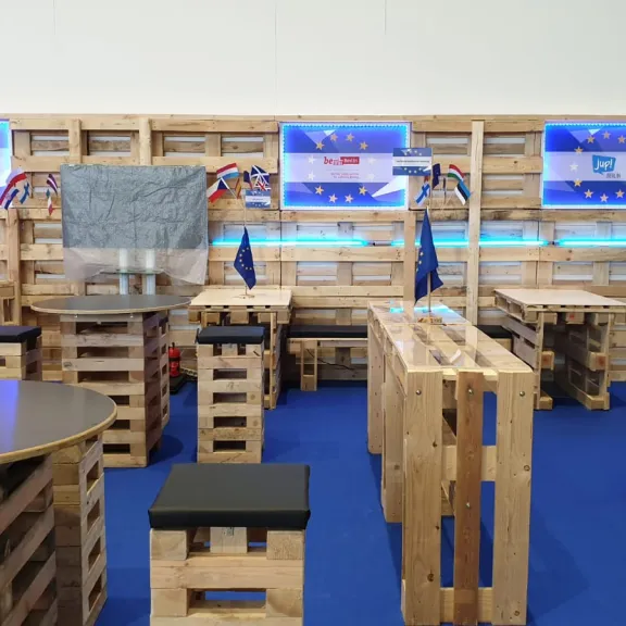 Raum mit Möbeln aus Paletten, EU-Flaggen und Bildschirmen, blauer Teppichboden.