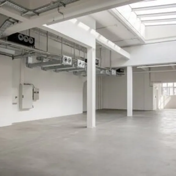 Leere, helle Industriehalle mit Betonboden und großen Fenstern.
