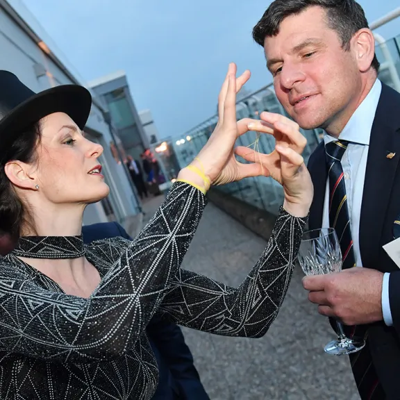 Frau mit Zylinder gestikuliert vor Mann im Anzug auf einer Dachterrasse.