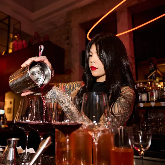 Frau hinter Bar gießt roten Cocktail in Gläser, stimmungsvolles Licht.