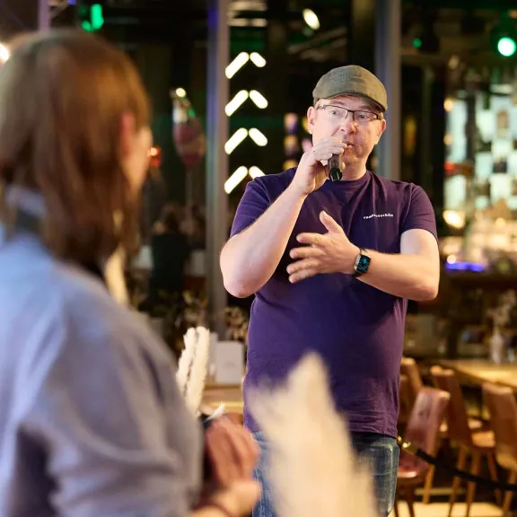 Mann mit Mikrofon spricht in einem Restaurant, eine Person im Vordergrund hört zu.