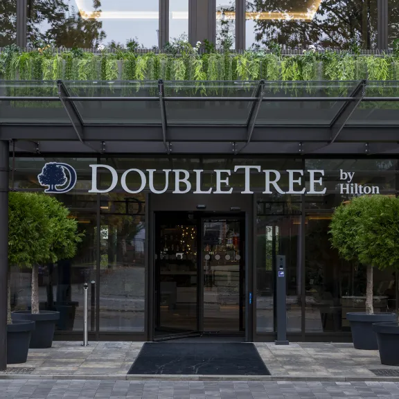 Hoteleingang mit DoubleTree by Hilton Logo, Glasdach und zwei großen Pflanzen.