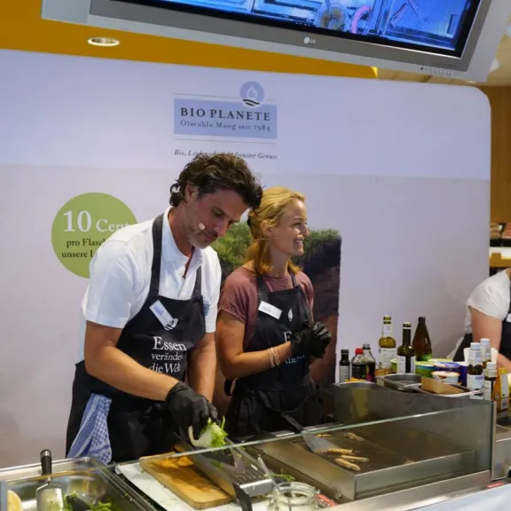 Zwei Personen kochen gemeinsam an einem Messestand mit Kochutensilien und Zutaten.