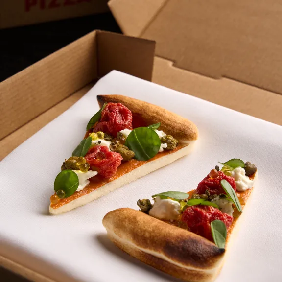 Zwei Pizzastücke mit Tomaten, Basilikum und Käse in einem offenen Pizzakarton.