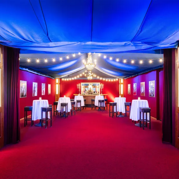 Festlich beleuchteter kleiner Saal mit Stehtischen, rotem Teppich und blauer Decke.