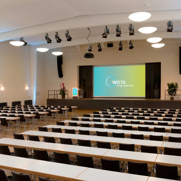 Großer, heller Konferenzsaal mit leeren Tischen und Leinwand auf der Bühne.