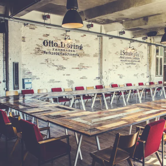 Großer, leerer Konferenzraum mit langen Holztischen und roten Stühlen, industrieller Stil.