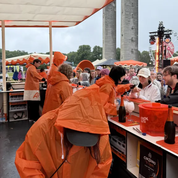 Menschen in orangefarbenen Regenponchos bedienen Kunden an einem Getränkestand im Freien.