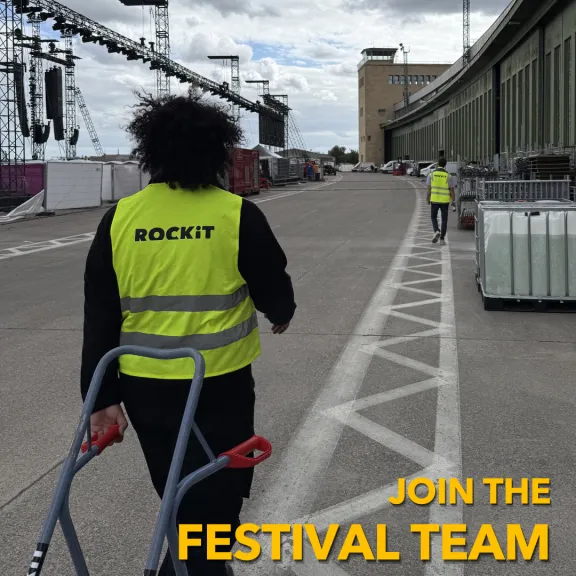 Zwei Personen mit gelben Westen auf einem Festivalgelände, eine zieht einen Wagen.