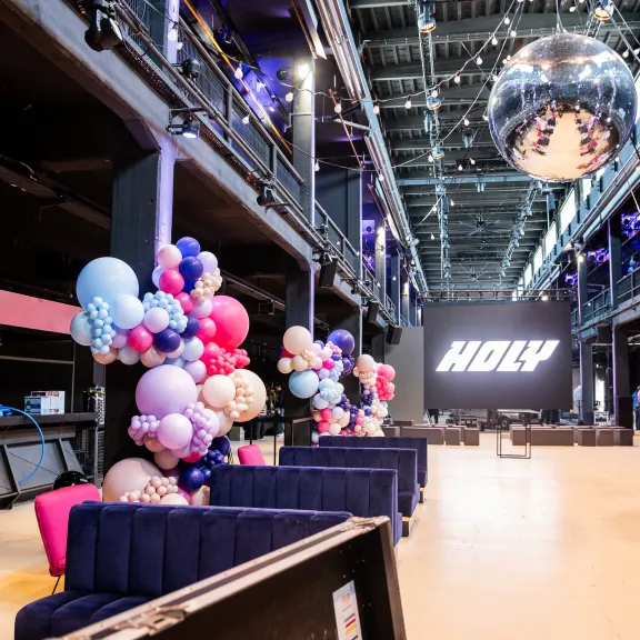 Große Eventhalle mit bunten Ballons, Sofas und einer Diskokugel an der Decke.