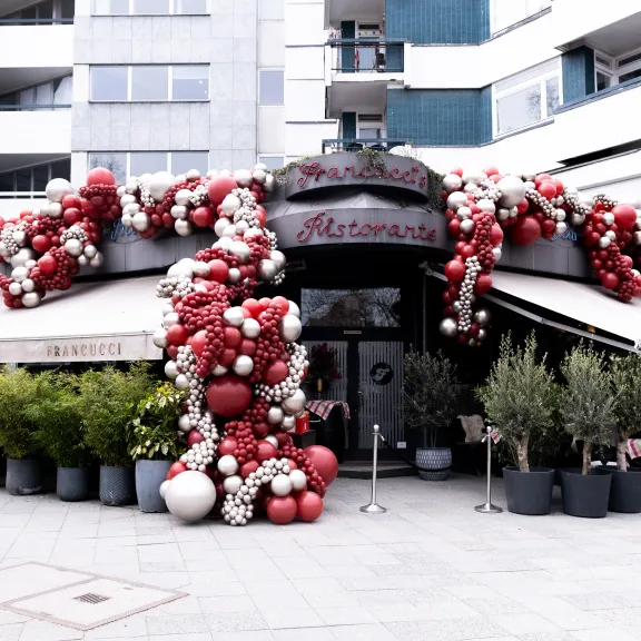 Restaurantfassade mit roter und weißer Luftballondekoration, umgeben von Pflanzen in Töpfen.