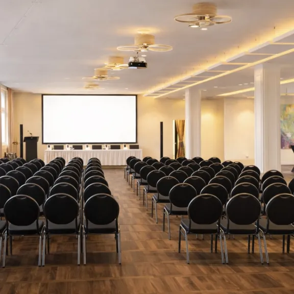Großer heller Konferenzraum mit vielen Stuhlreihen und einer Leinwand vorne.