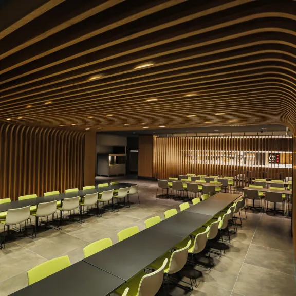 Modernes Restaurant mit langen Tischen, grünen Stühlen und Holzdecke.